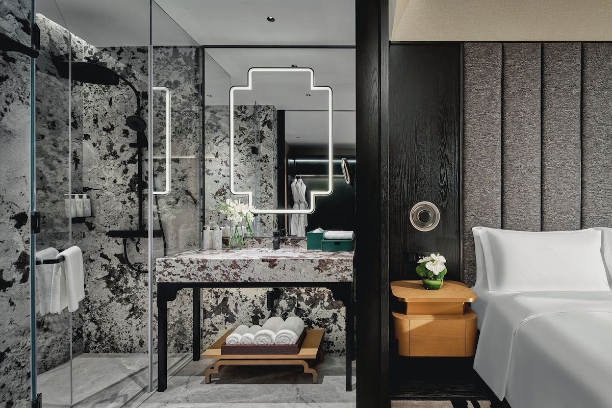 Un baño moderno con tocador de mármol, cabina de ducha de vidrio y mesita de noche de madera con una silla blanca.