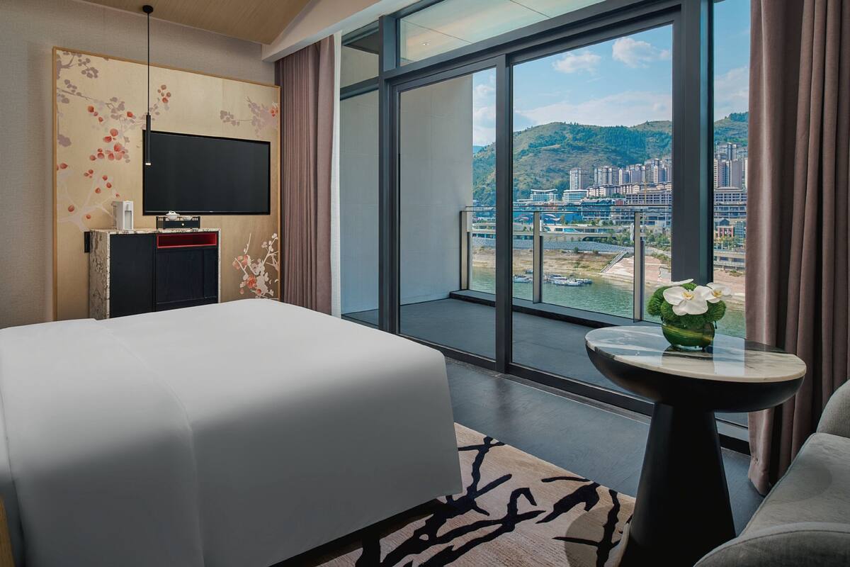 A hotel room with a large bed, a TV on the wall, a small table with a vase, and a balcony with a city view.