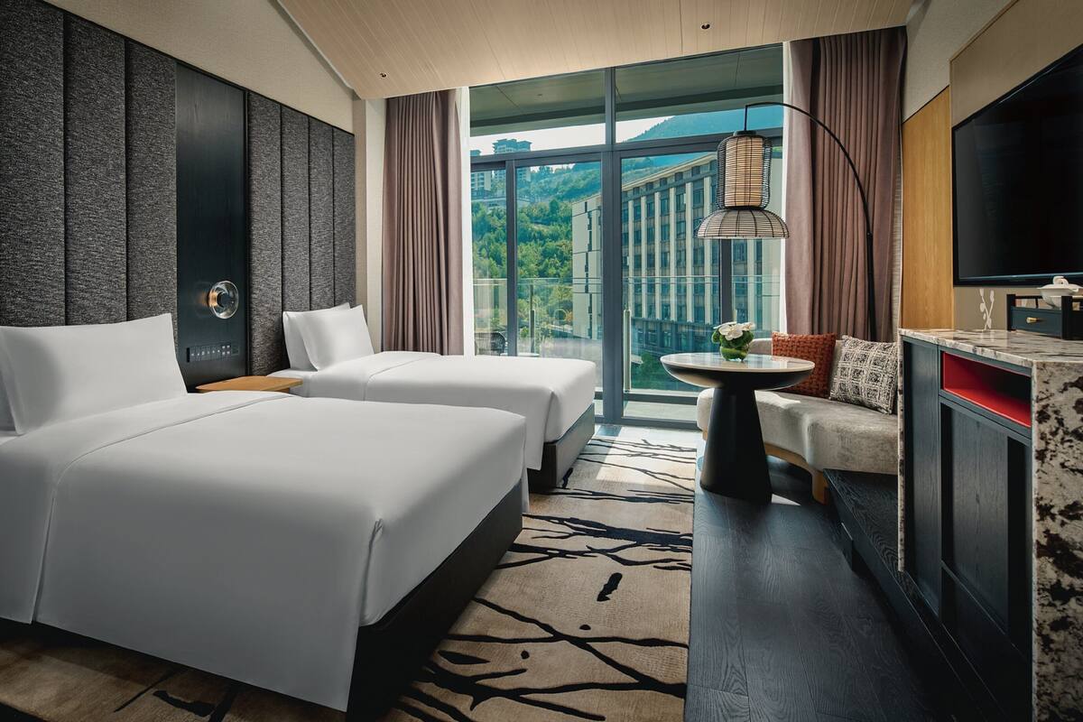 A hotel room with two beds, a sofa, a small table, a TV, and a large window with a view of buildings and greenery.