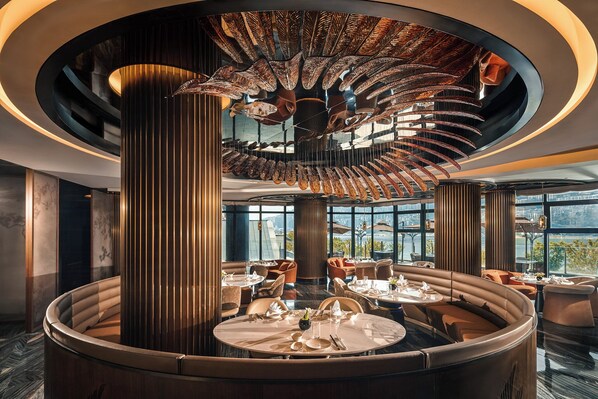 Un restaurante moderno con una zona de estar circular central, amplios ventanales con vistas a la ciudad y un diseño de techo singular.