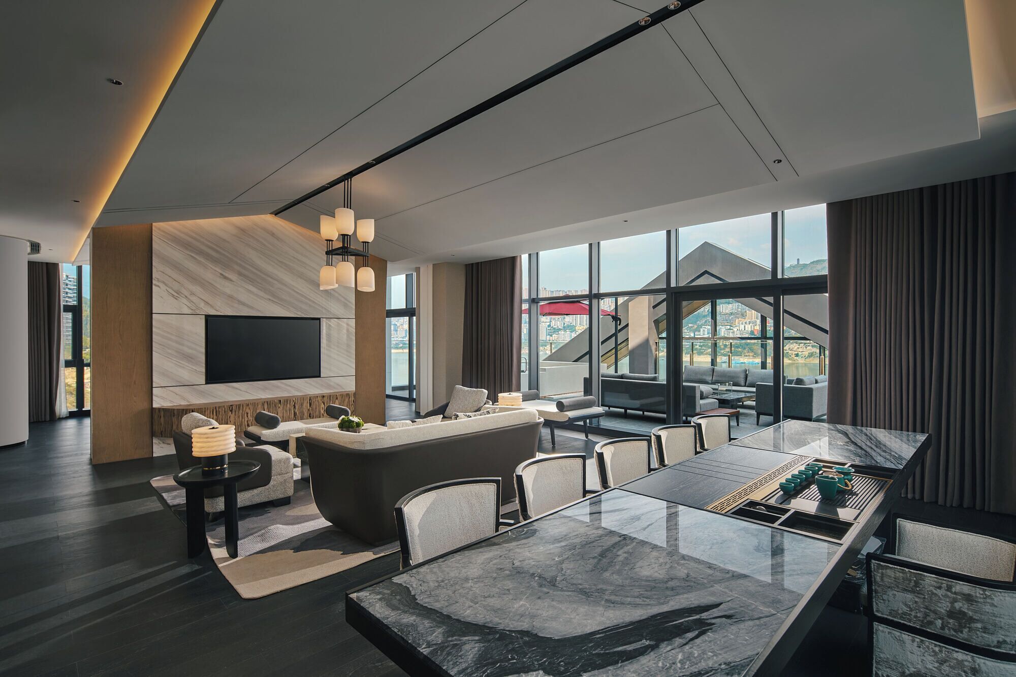 A modern hotel room with a large dining table, comfortable seating, a TV mounted on the wall, and a view of the city through large windows.