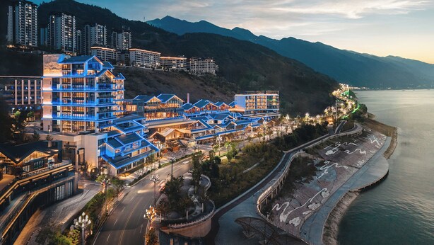 Un hotel moderno con varios pisos, un paseo marítimo y montañas al fondo.