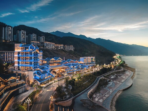 Un hotel moderno con varios pisos, un paseo marítimo y montañas al fondo.