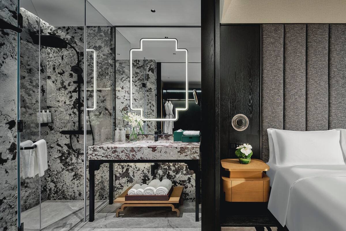 A modern bathroom with a marble vanity, a glass shower enclosure, and a wooden nightstand with a white chair.