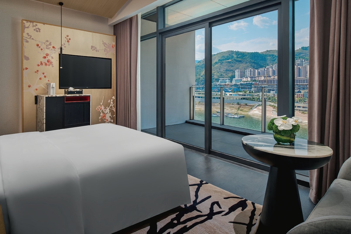 Habitación de hotel con una cama grande, un televisor en la pared, una mesita con un jarrón y un balcón con vistas a la ciudad.