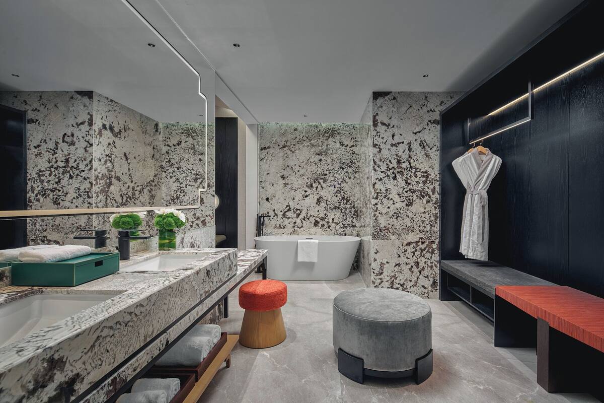 A modern bathroom with a freestanding bathtub, a vanity with a marble countertop, and a bench with a red cushion.