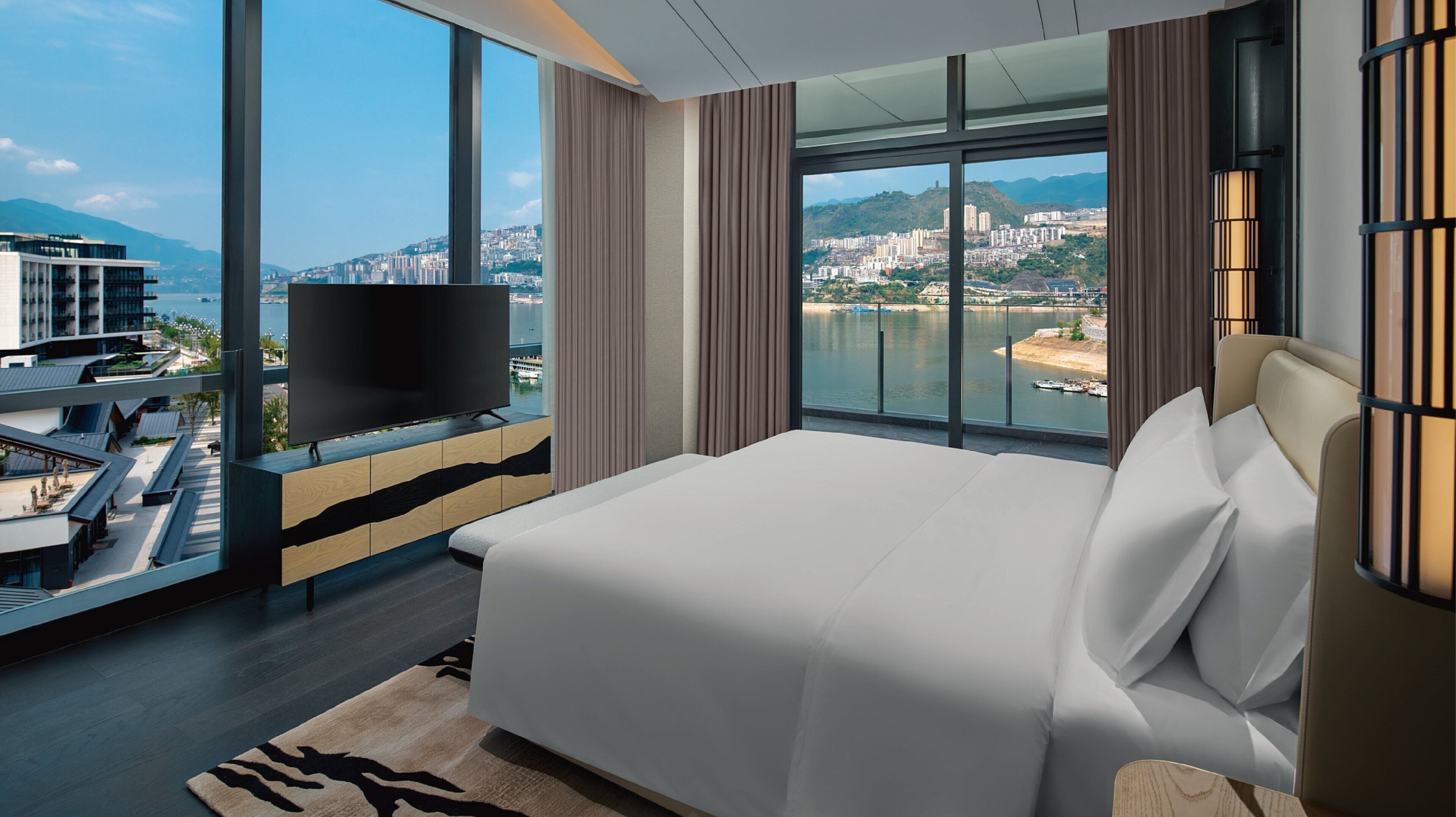A modern hotel room with a large bed, a TV, and a view of a city and water.