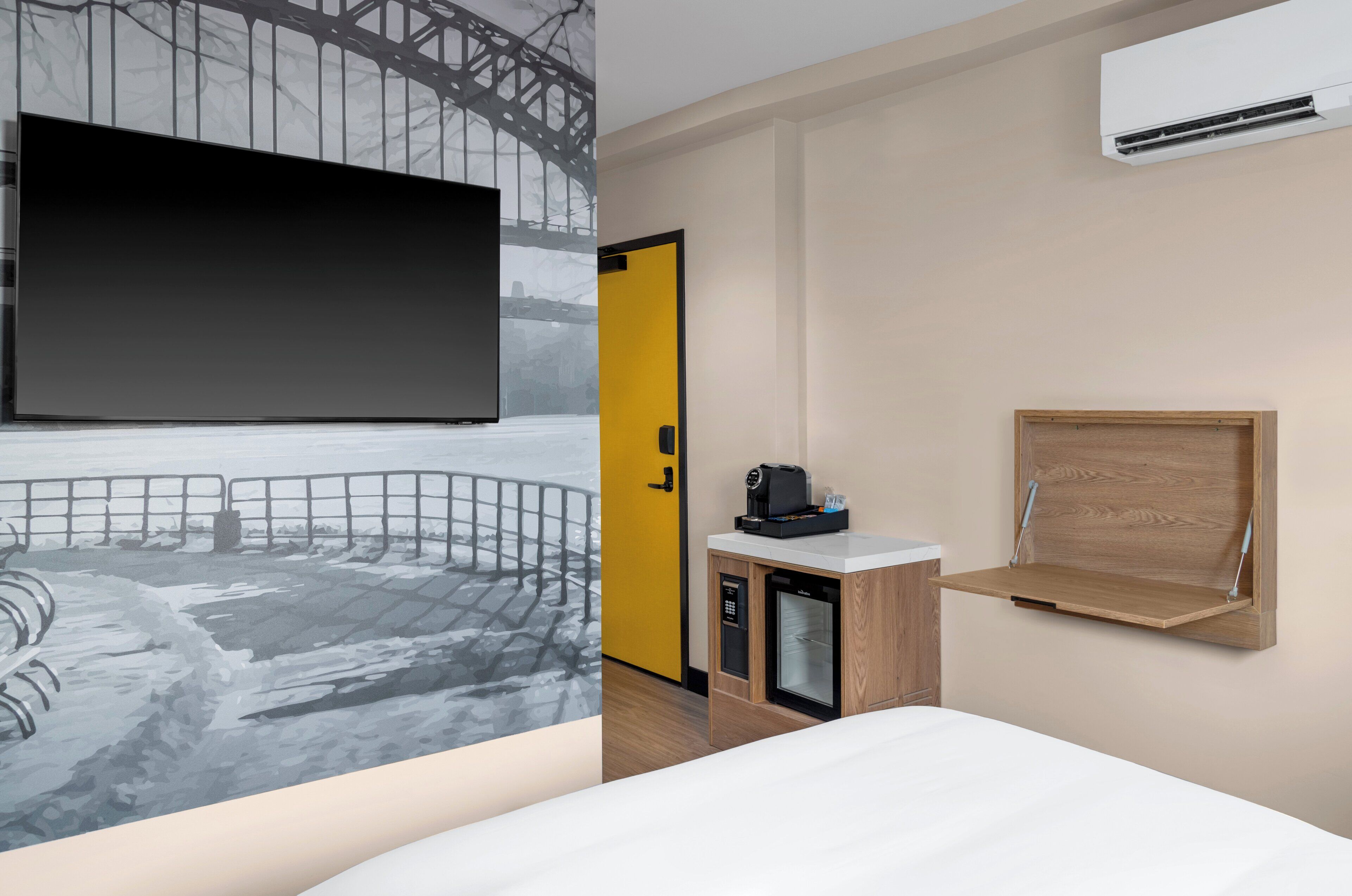 A hotel room with a large flat-screen TV, a wooden cabinet with a coffee machine, a small refrigerator, and a wooden shelf.