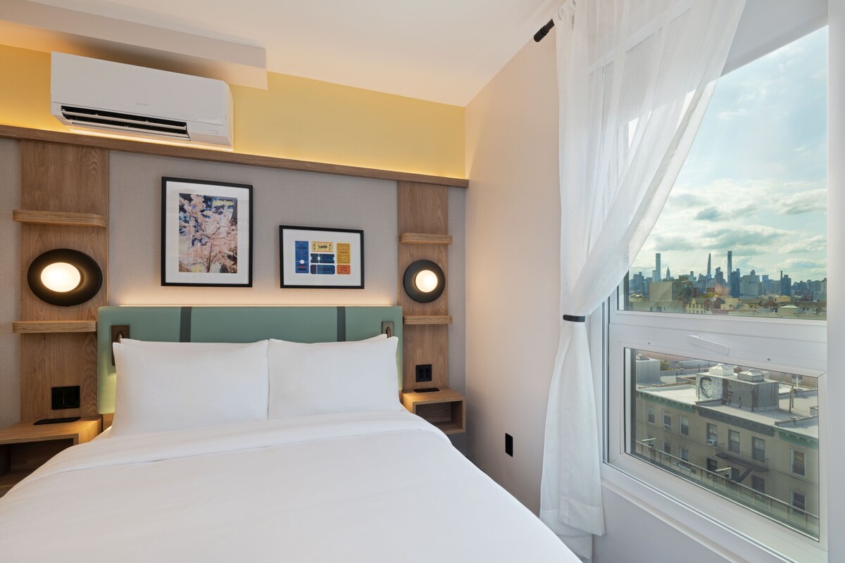 Habitación de hotel con cama, vista a la ciudad, aire acondicionado y cuadros en la pared.