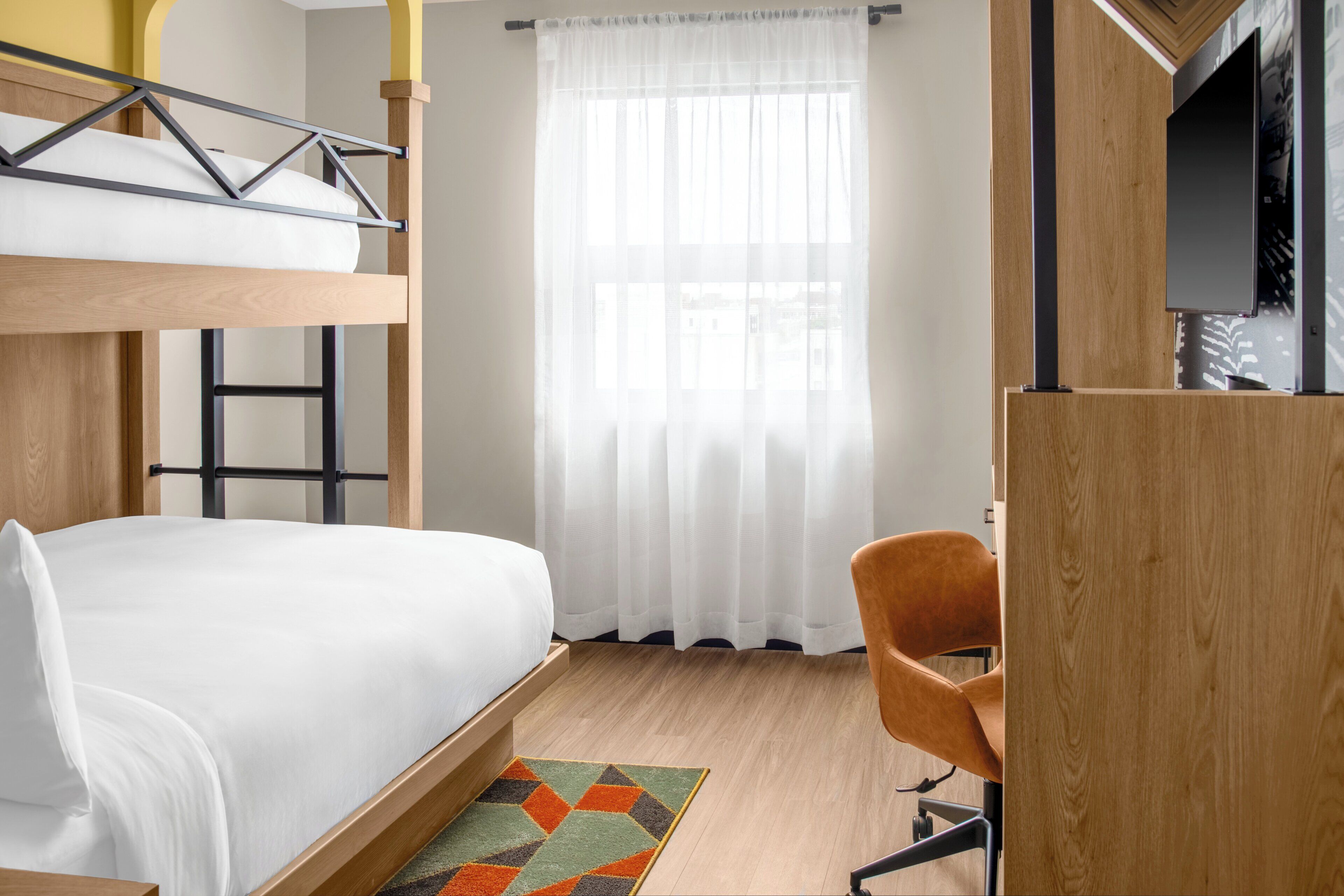 A bunk bed with a wooden frame and white bedding, a desk with a chair, a large window with sheer curtains, and a television on a wooden stand.