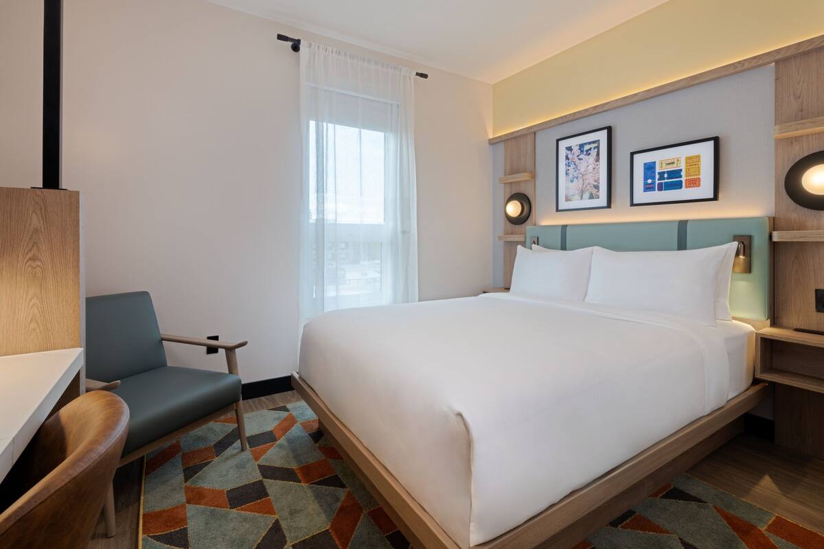 Une chambre d’hôtel comprenant un lit, un bureau, une chaise, une fenêtre avec des rideaux légers et des œuvres d’art accrochées au mur.