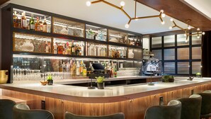Zona de bar con una barra curva, múltiples estantes con botellas de licores, una caja registradora y taburetes de bar tapizados en verde.