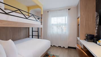 A bunk bed with a white mattress and pillows, a small desk with a coffee maker, a window with white curtains, and a geometric rug on the floor.