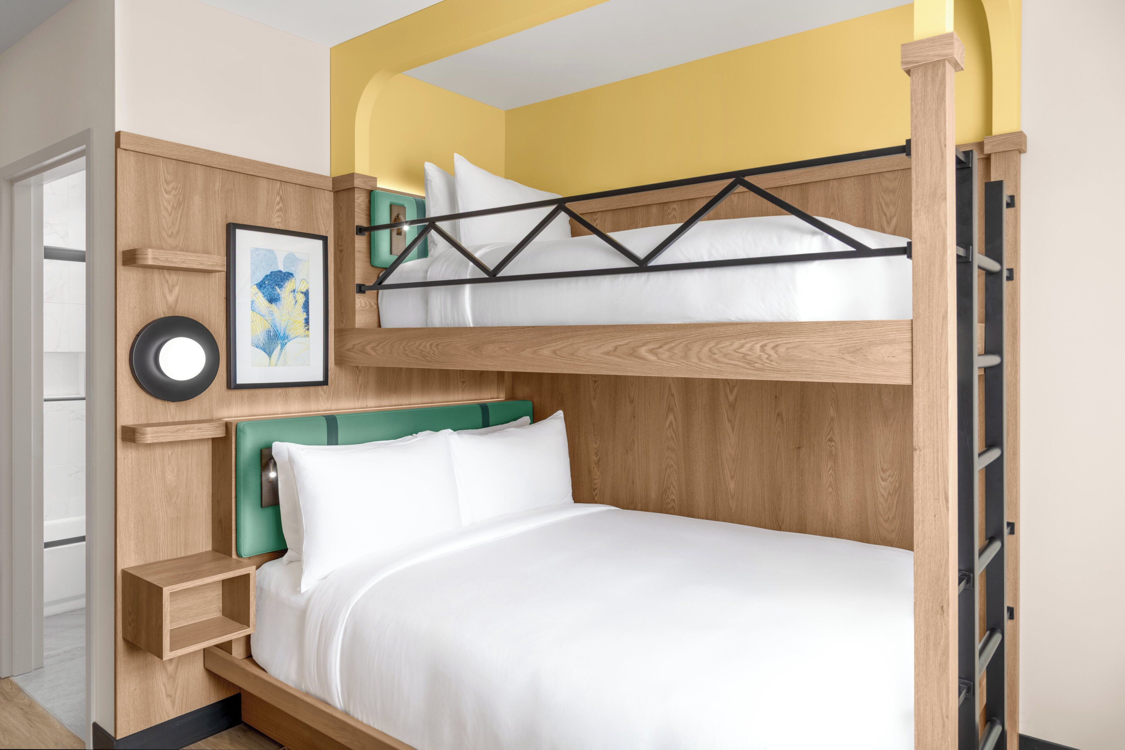 Una cama litera con estructura de madera y ropa de cama blanca, una mesita de noche con cabecera verde, y un cuadro en la pared.