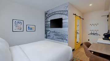 A hotel room with a bed, a desk, a chair, a mirror, a coat rack, and a wall with a large picture and two smaller framed pictures.