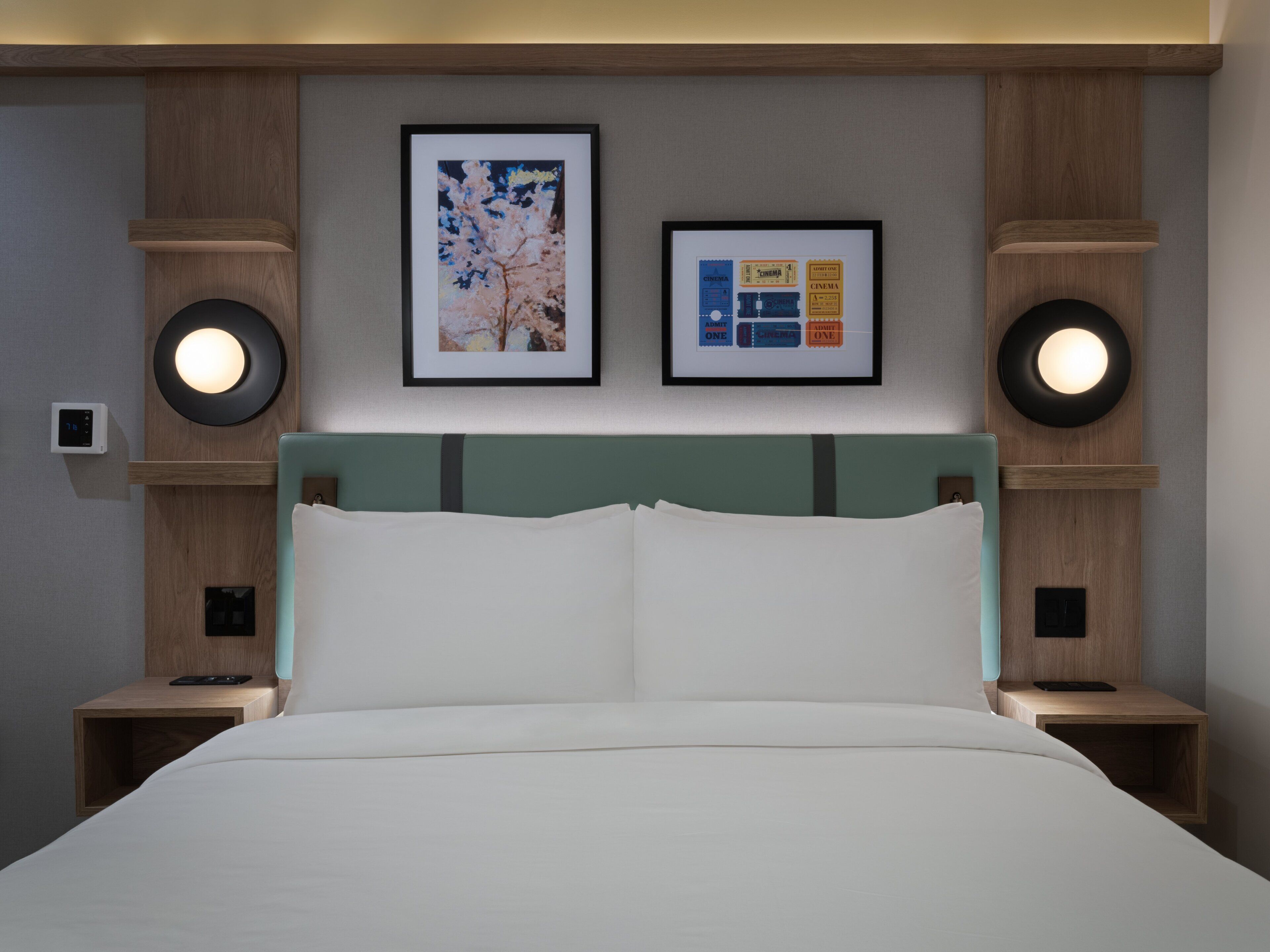 Ein Hotelzimmer mit einem Bett, einem Kopfteil, zwei Nachttischen, einer Wandleuchte und zwei gerahmten Bildern.