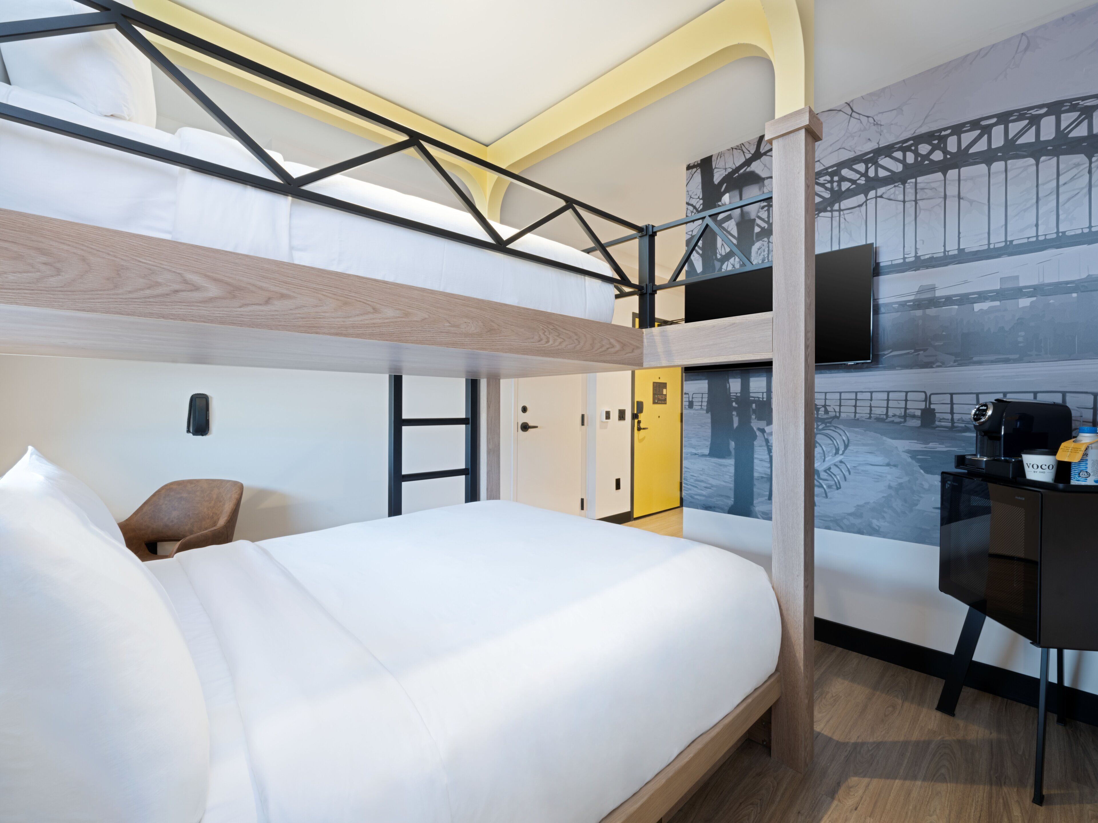 A modern hotel room with a bunk bed, a desk with a coffee maker, and a mural of a bridge.
