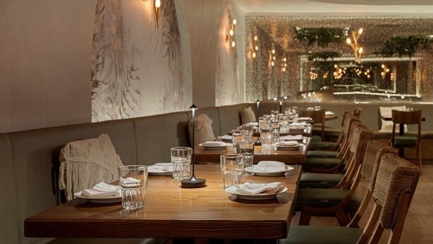 Un restaurante con una larga mesa de madera dispuesta para comer, sillas tapizadas y una pared decorativa con una planta.