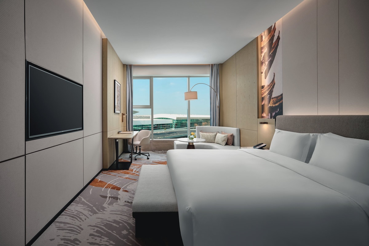 A modern hotel room with a large bed, a desk, a chair, a TV, and a view of the city.