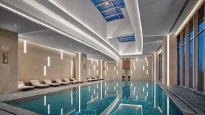 A modern indoor swimming pool with a clear blue water surface, surrounded by lounge chairs and a high ceiling with skylights.