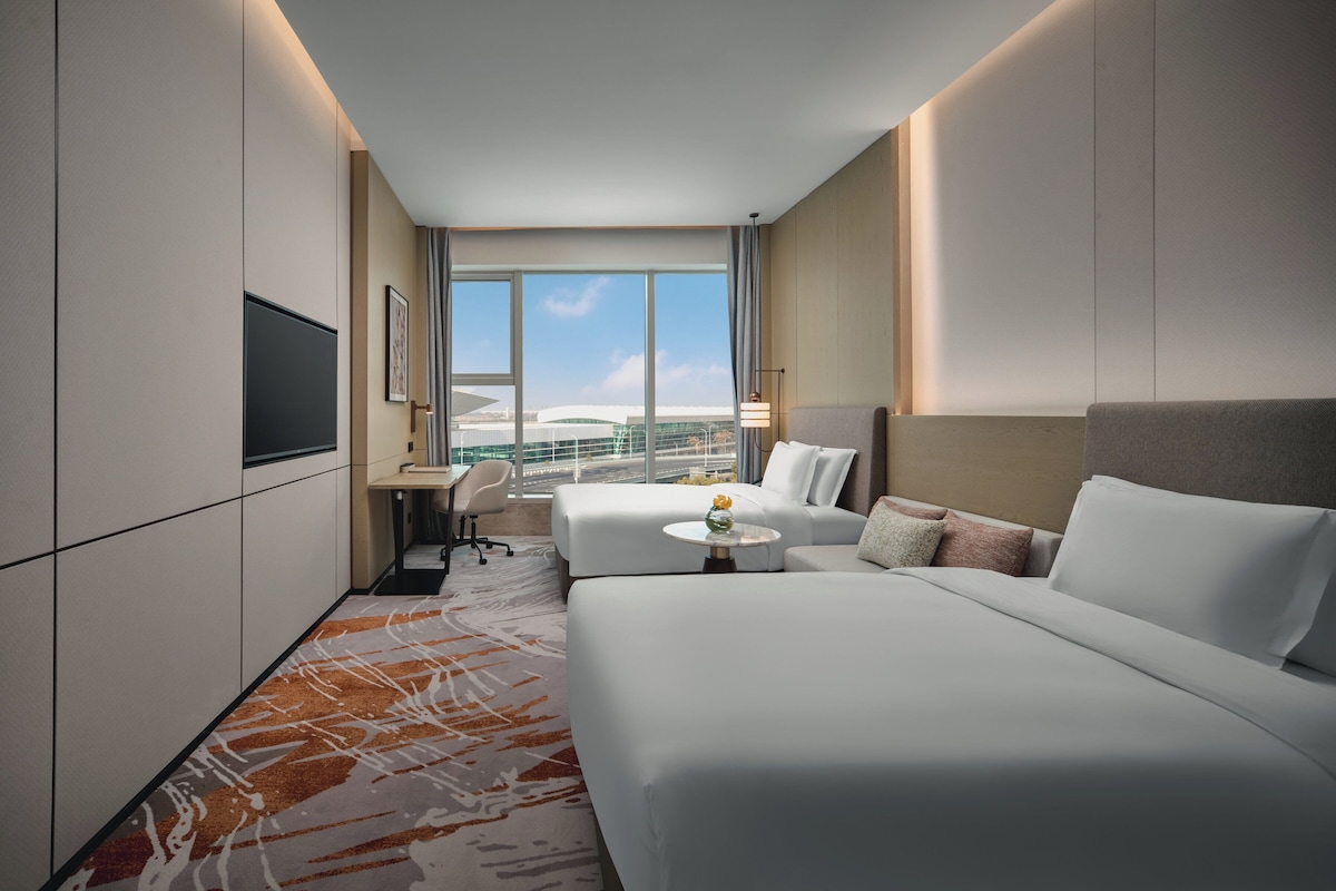 A hotel room with two beds, a desk, a chair, and a large window offering a view of a building.