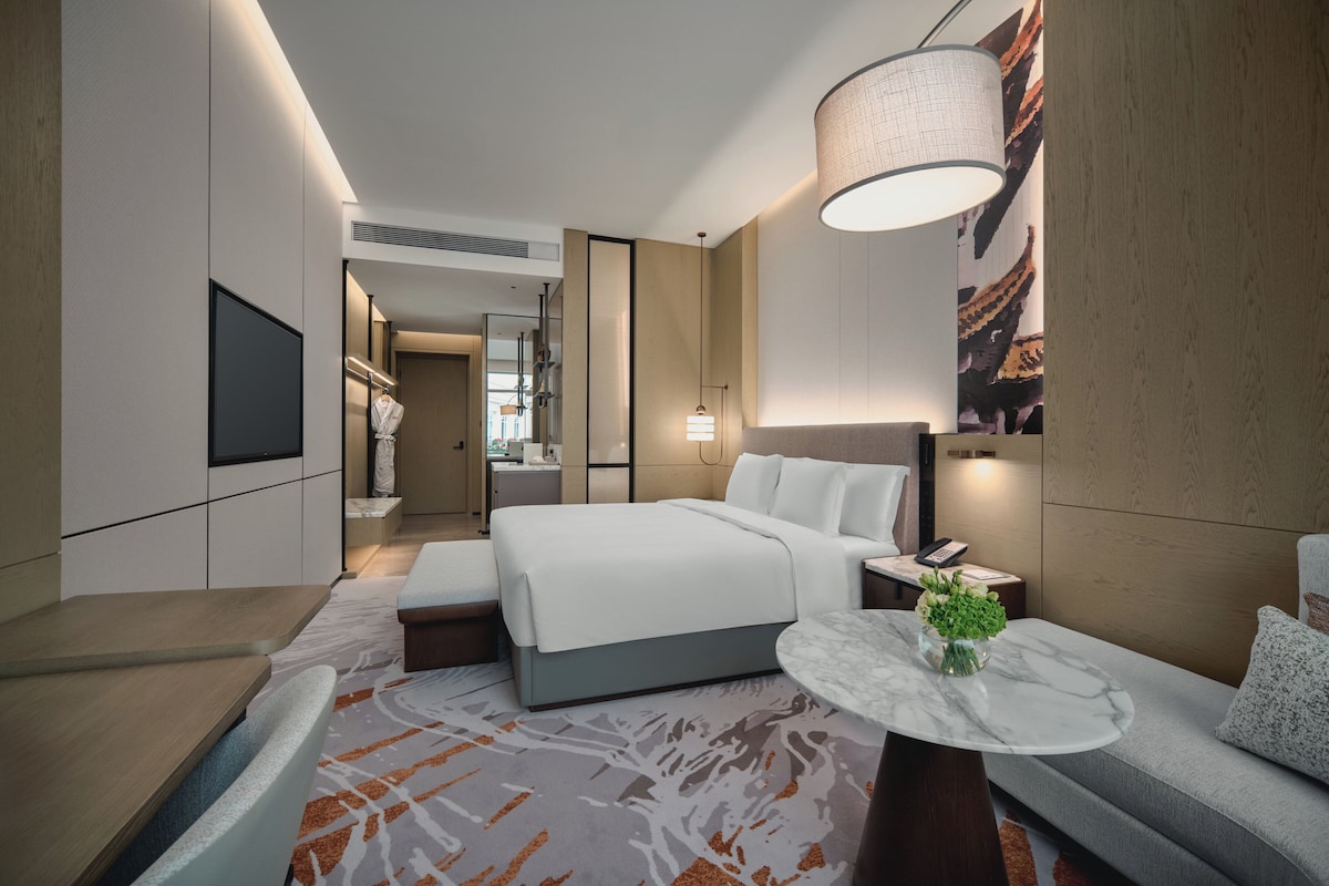 A modern hotel room with a large bed, a seating area with a small table, a TV mounted on the wall, and a large window with a view.