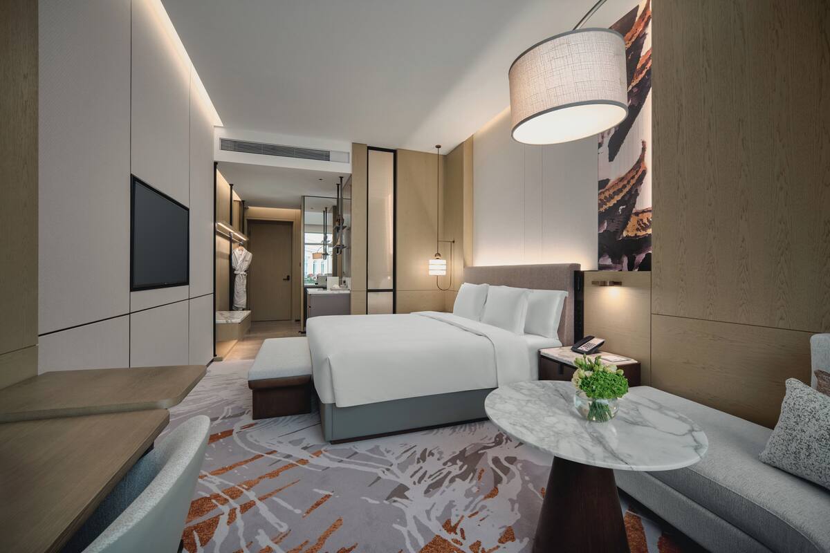 A modern hotel room with a large bed, a seating area with a small table, a TV mounted on the wall, and a large window with a view.