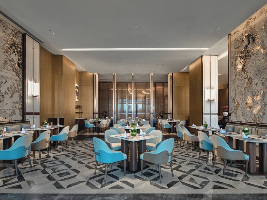 Um hall de hotel moderno com várias mesas e cadeiras para refeições, paredes de mármore e piso com padrão geométrico.