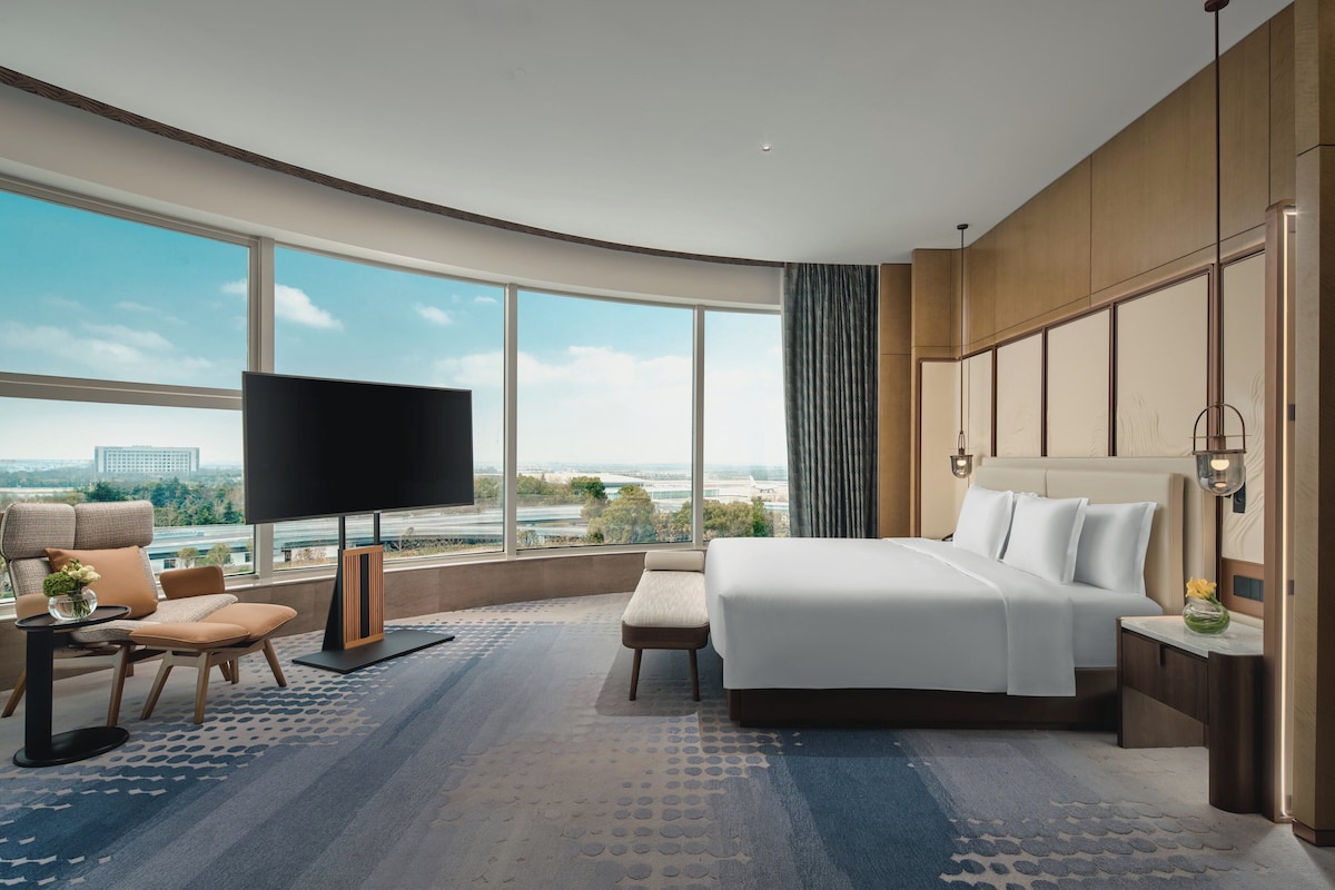 Una habitación de hotel moderna con una cama grande, un televisor de pantalla plana, una zona de estar con una silla y una mesa, y vistas de la ciudad a través de amplias ventanas.