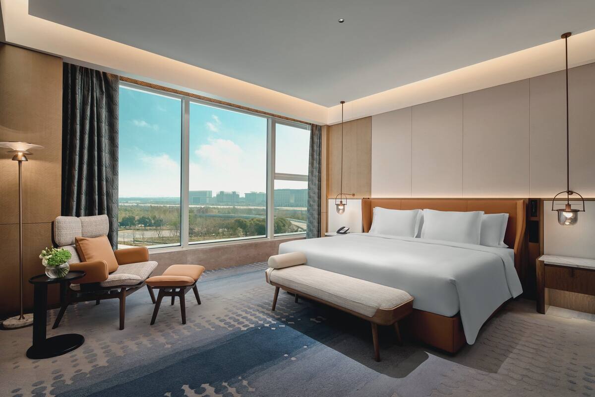 A modern hotel room with a large bed, a seating area with a sofa and armchair, a small table with a plant, and a large window offering a city view.