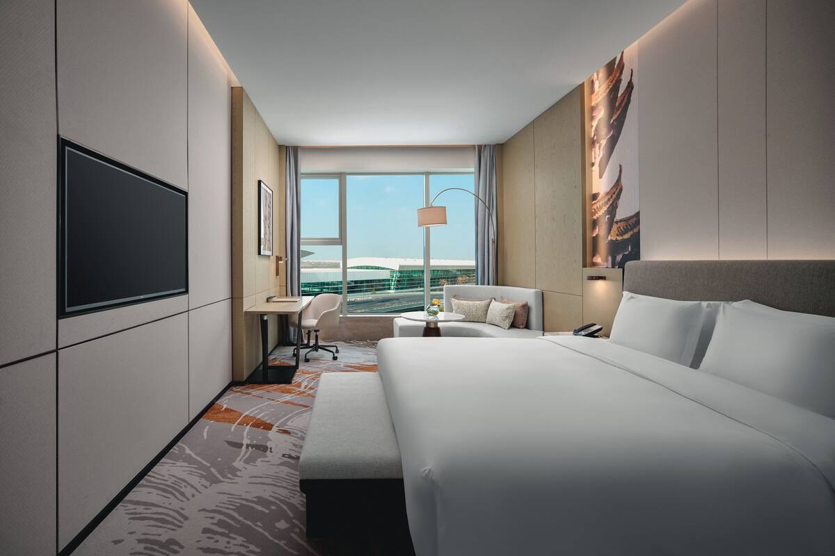A modern hotel room with a large bed, a desk, a chair, a TV, and a view of the city.