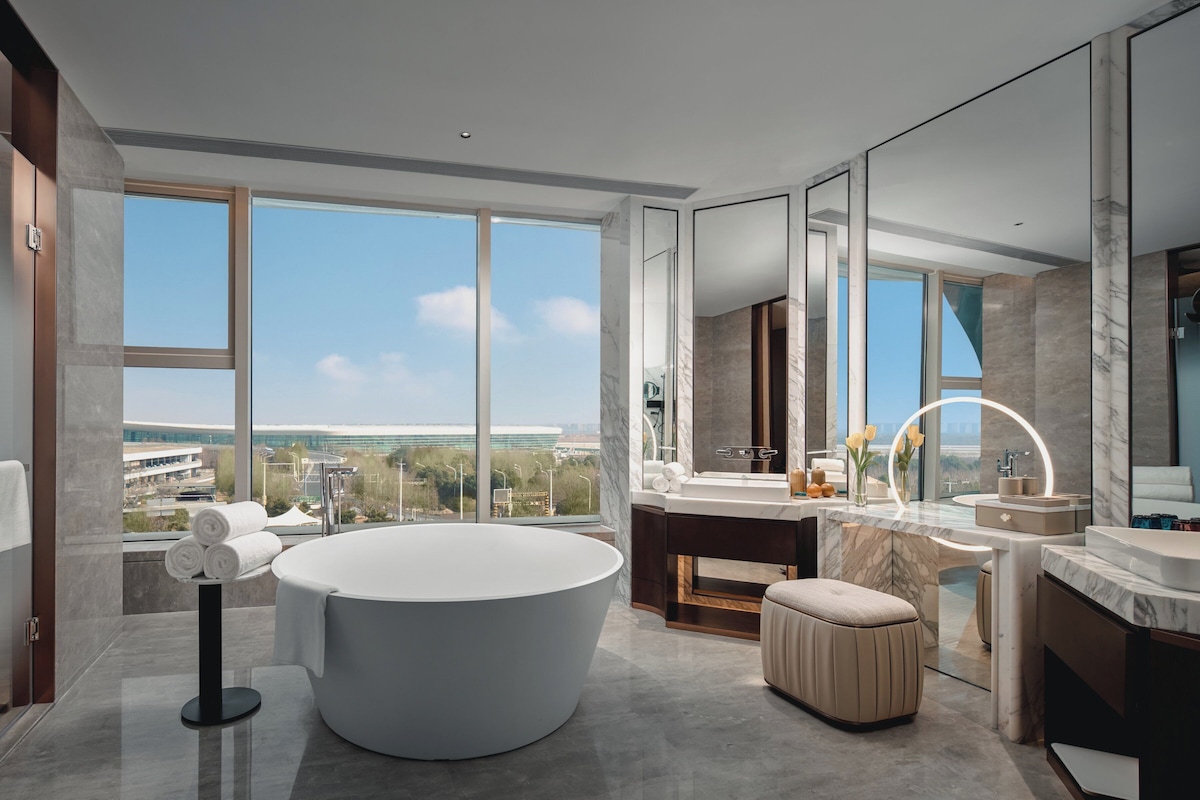A modern bathroom with a large bathtub, a vanity with a mirror, and a view of the cityscape through large windows.
