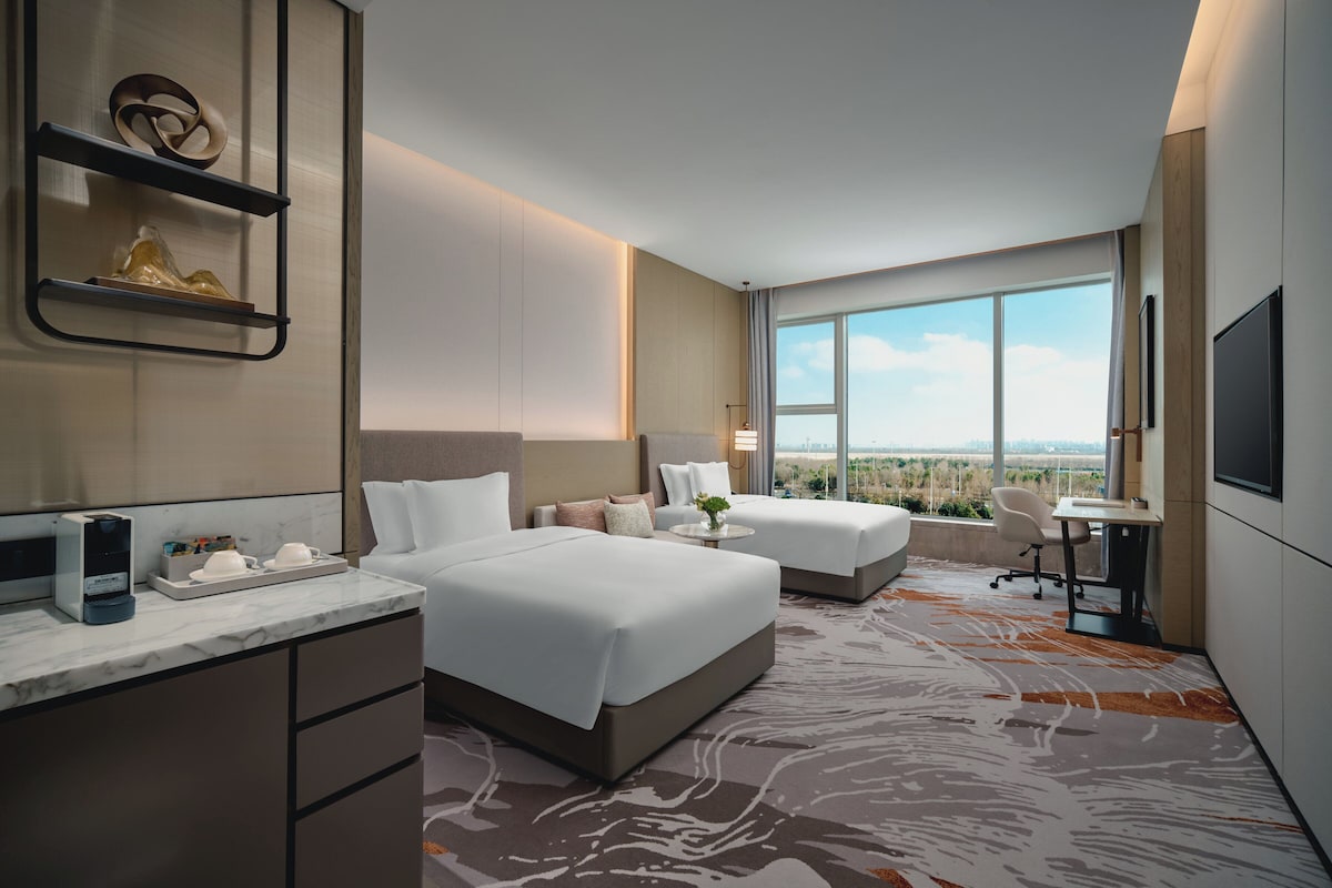 A modern hotel room with two beds, a desk, and a large window offering a city view.