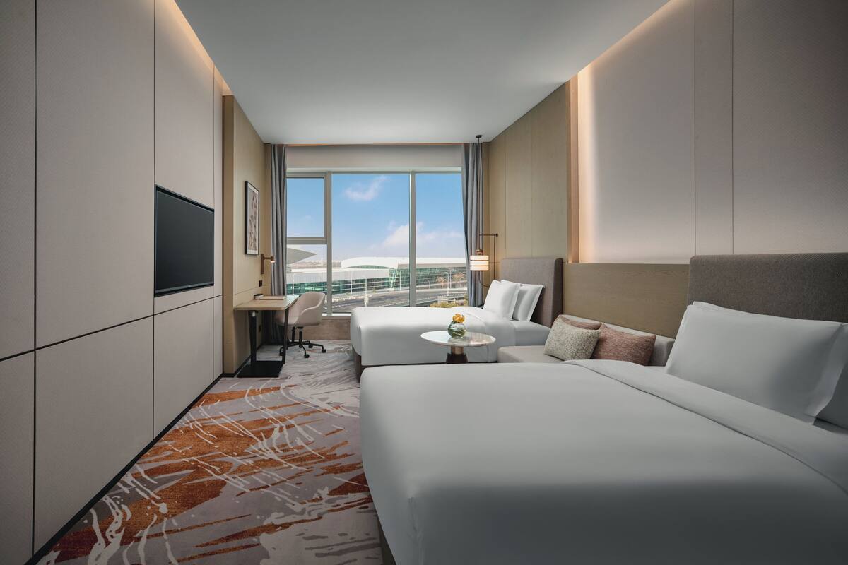 A hotel room with two beds, a desk, a chair, and a large window offering a view of a building.