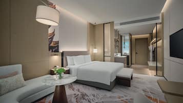 Ein modernes Hotelzimmer mit einem großen Bett, einer Sitzecke mit einer Couch und einem kleinen Tisch, einem Badezimmer mit Spiegel und Dusche sowie einem an der Wand befestigten Flachbildfernseher.