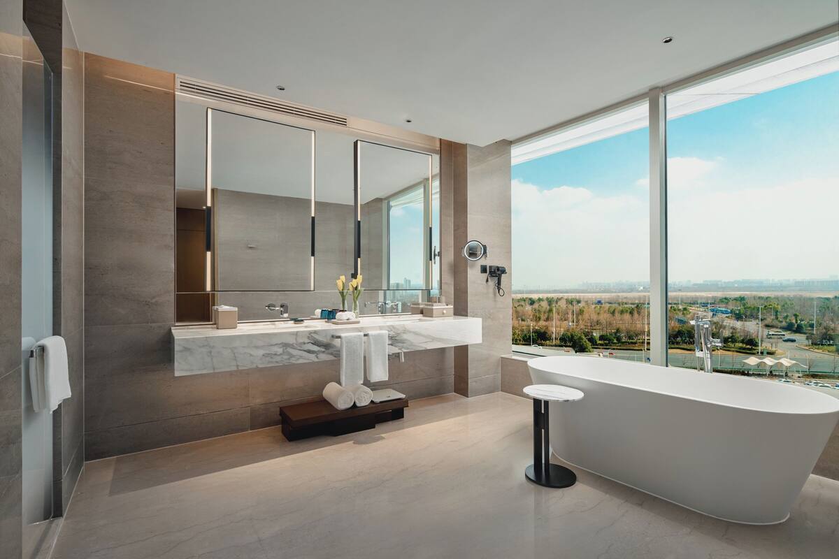 A modern bathroom with a large bathtub, a double vanity with a large mirror, and a view of a cityscape through large windows.