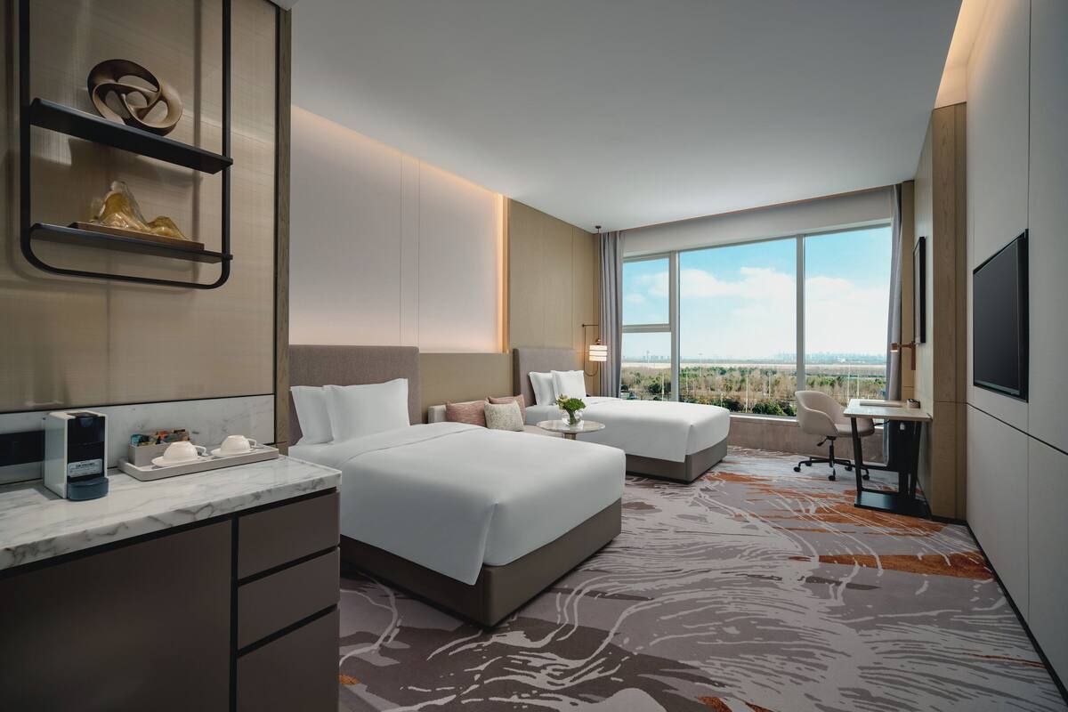 A modern hotel room with two beds, a desk, and a large window offering a city view.