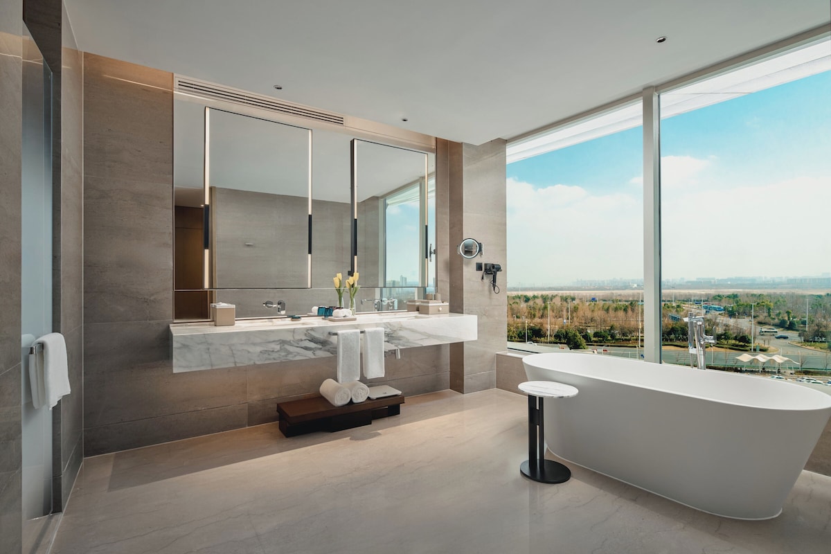 A modern bathroom with a large bathtub, a double vanity with a large mirror, and a view of a cityscape through large windows.