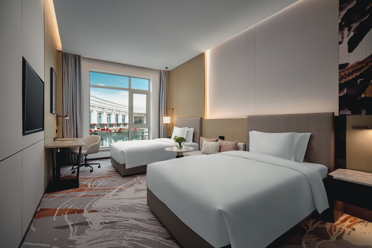 Habitación de hotel con dos camas, un escritorio, una silla, una mesita con un florero y un amplio ventanal con vistas a un edificio y una piscina.