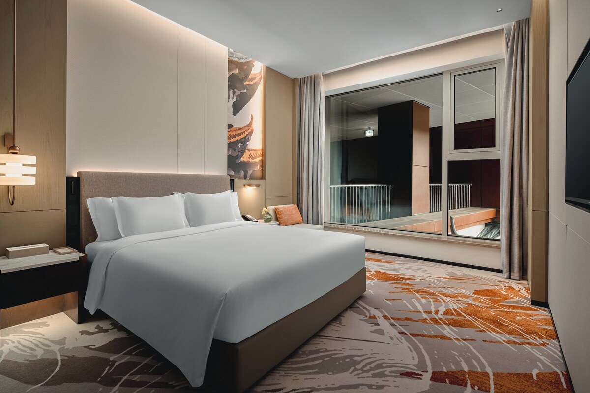 A modern hotel room with a large bed, a nightstand, a wall-mounted lamp, a large window with a balcony, and a view of the cityscape.