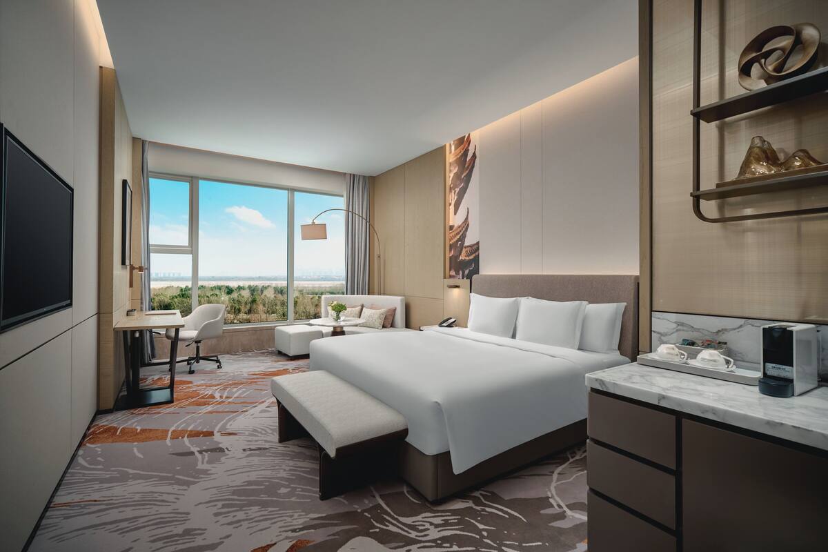 A modern hotel room with a large bed, a desk, a chair, a bench, a TV, and a coffee machine.