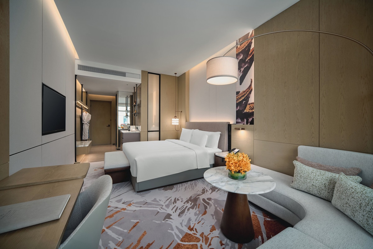 A modern hotel room with a bed, a sofa, a dining table, and a TV.