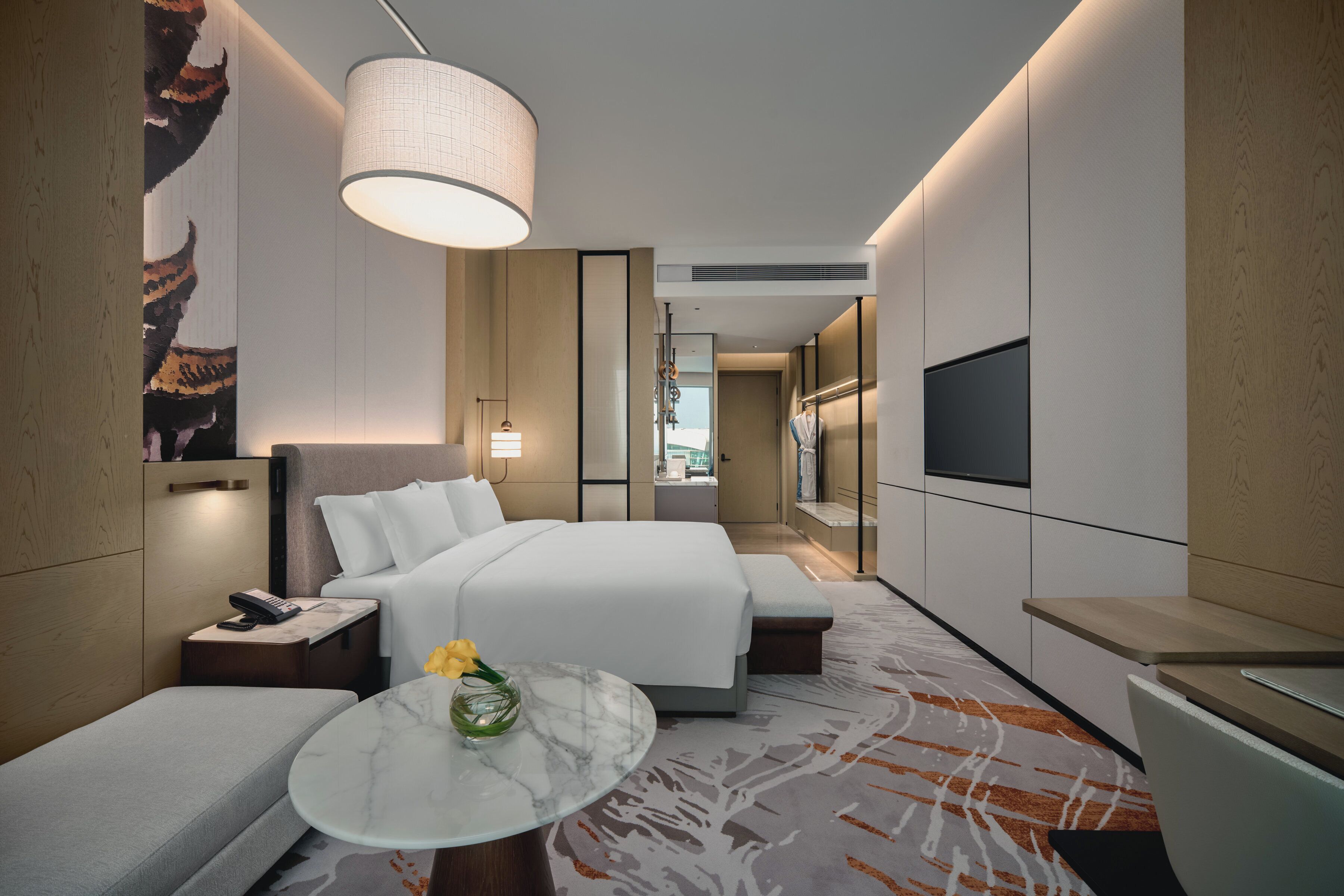 A modern hotel room with a large bed, a small seating area, a TV, and a bathroom visible in the background.