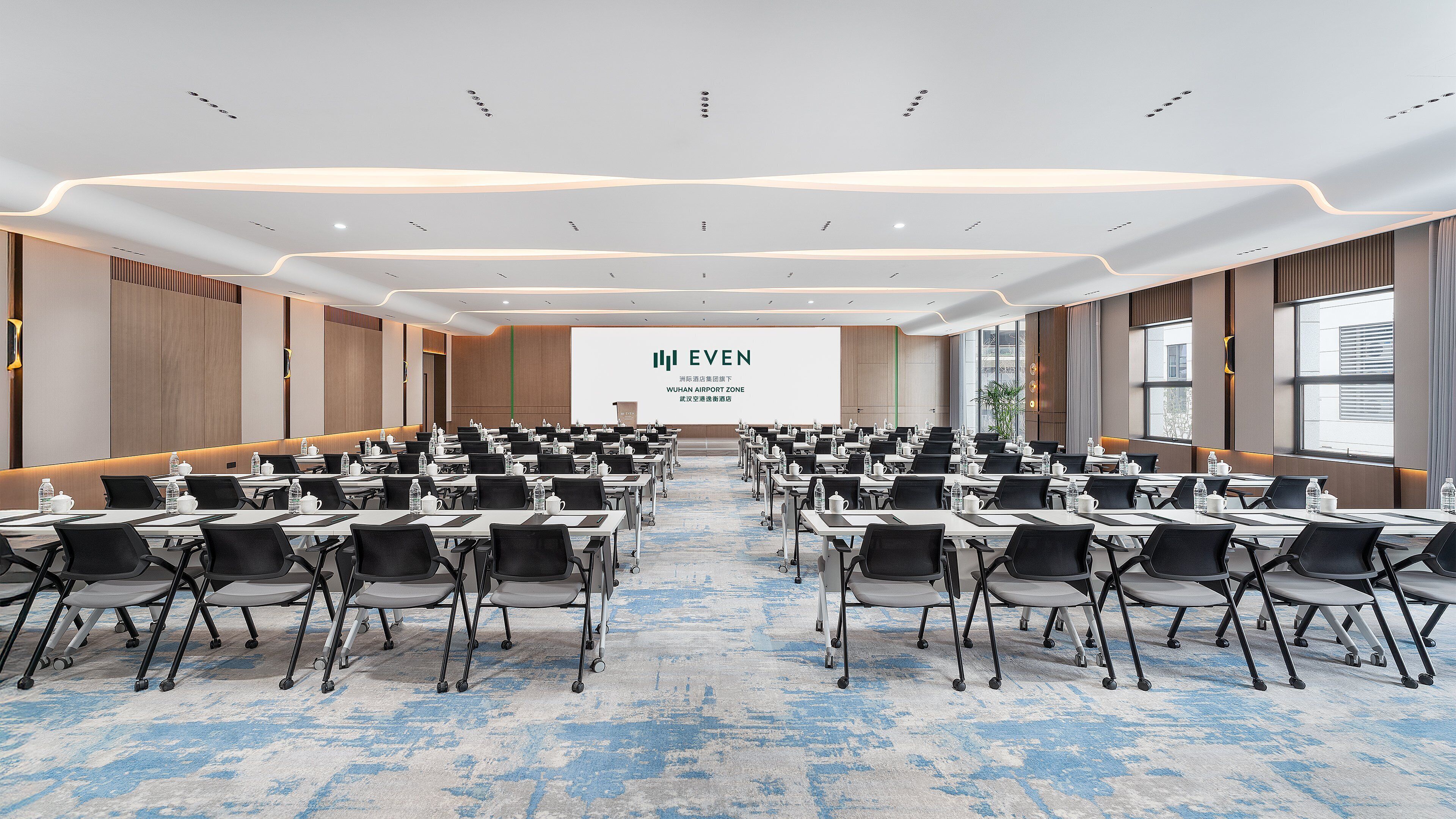 A large conference room with rows of tables and chairs, a presentation screen, and a view of the airport zone.