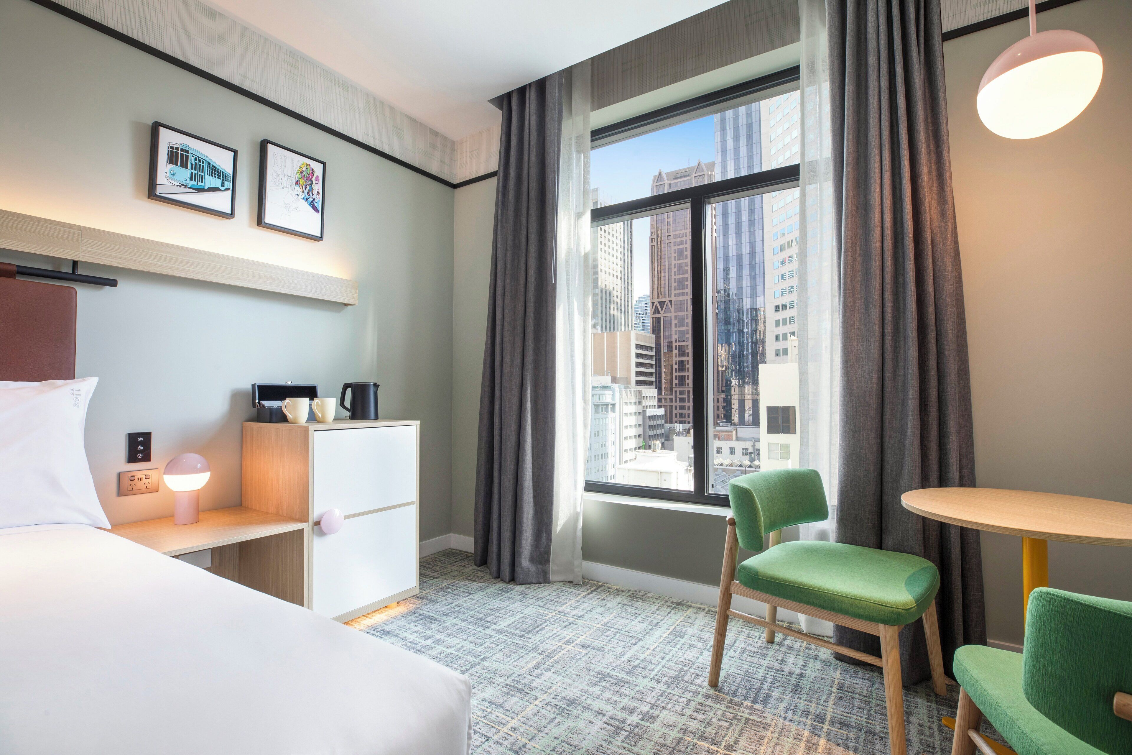 A hotel room with a bed, a desk with a lamp, a chair, a small table, and a window with curtains.