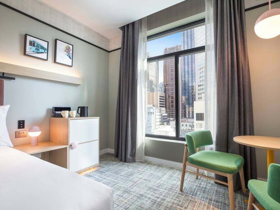 Hotelkamer met een bed, een bureau met lamp, een stoel, een tafeltje en een raam met gordijnen.