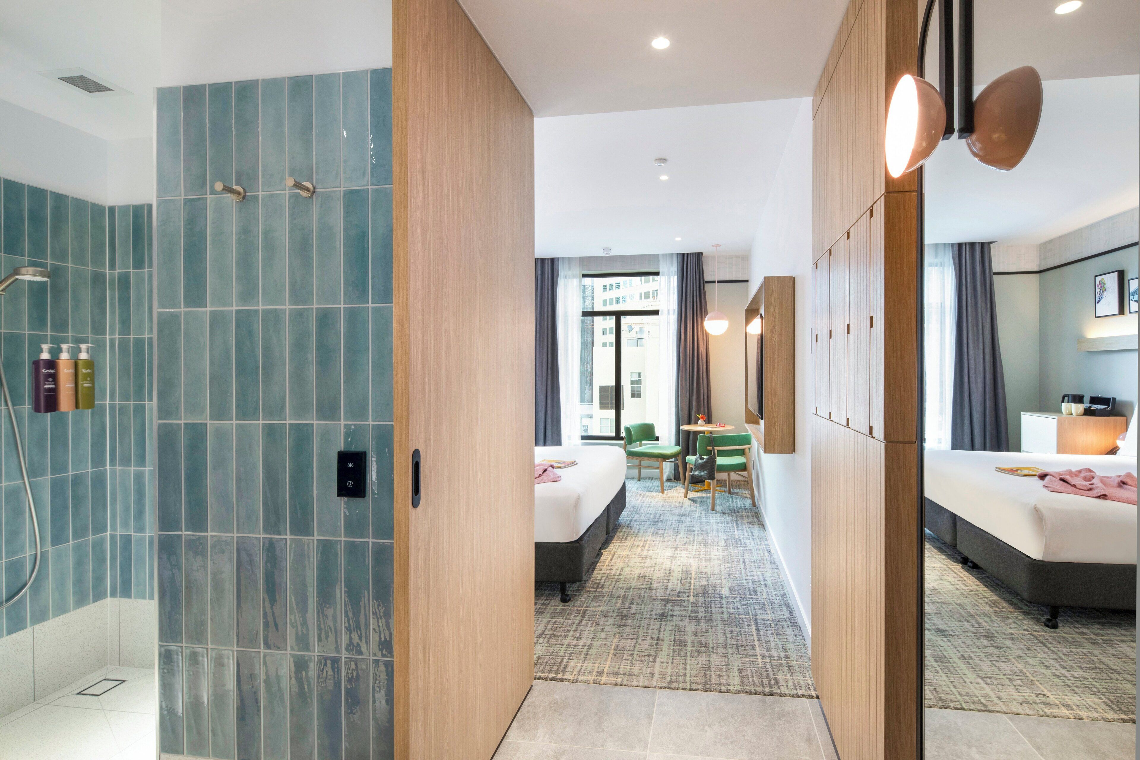 A hotel room with a shower, a bed, a desk, and a view of the city.