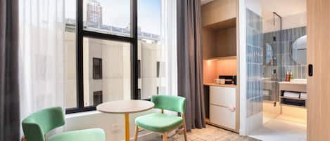 A hotel room with a large window, two green chairs, a small round table, a wooden cabinet, and a bathroom with a mirror and sink.