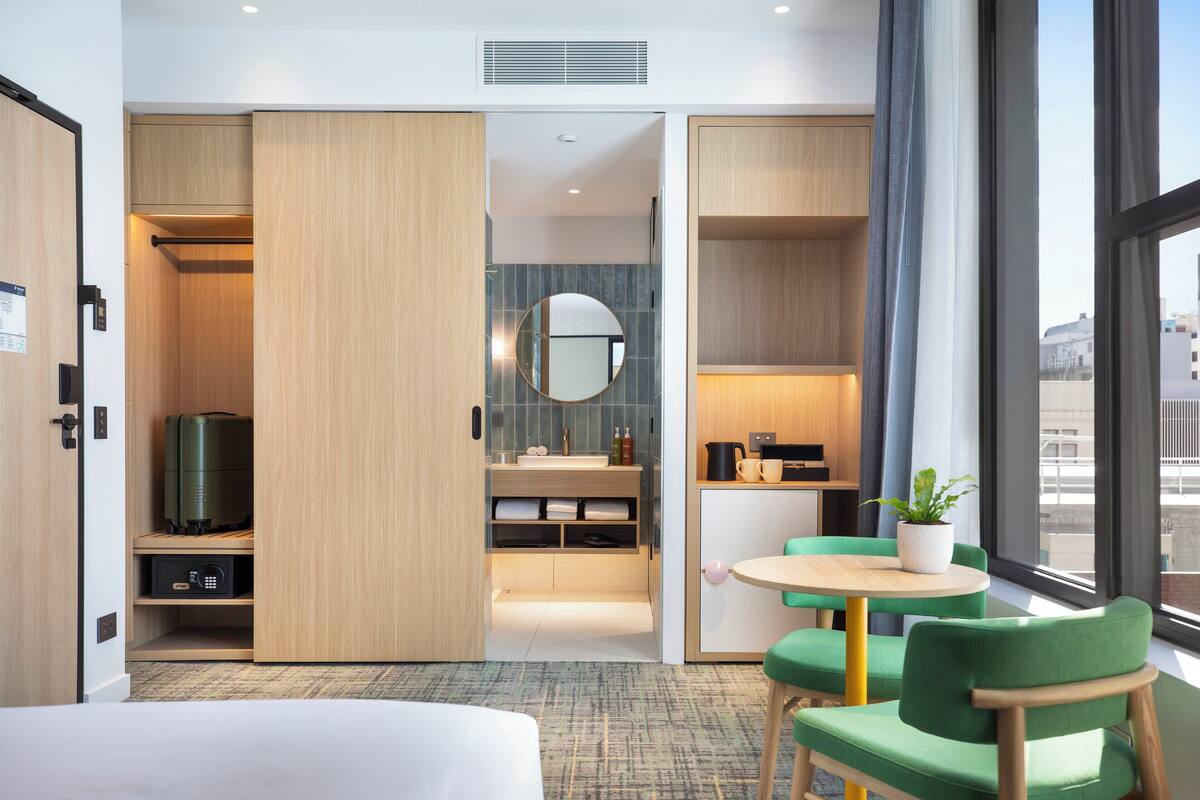 Une chambre d’hôtel avec un lit, un canapé vert, une petite table, un miroir, un lavabo et une vue sur la ville grâce à de grandes fenêtres.
