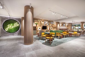 Een moderne hotellobby met een groen bord waarop 'Hello' staat, houten accenten en een zithoek met gele stoelen.
